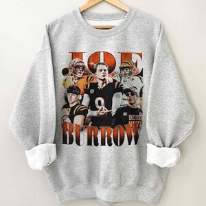 May include: A heather gray crewneck sweatshirt featuring a graphic print of football player Joe Burrow in various poses and outfits. The text JOE BURROW is printed in large orange and black block letters above and below the photographic images.