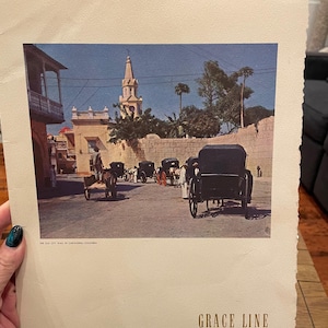 Könnte beinhalten: Ein alter Papierdruck mit einem Farbfoto von Pferdekutschen auf einer Straße neben einer Steinmauer und einem Kirchturm in Cartagena, Kolumbien. Der Text "THE OLD CITY WALL IN CARTAGENA, COLOMBIA" und "GRACE LINE" ist auf dem Papier aufgedruckt.