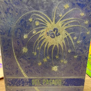 May include: A blue hardcover yearbook with a textured finish featuring a gold-colored firework graphic and the text '62' in the center. The bottom section includes a horizontal band with the text 'DEL PASADO' printed in a dark green font.