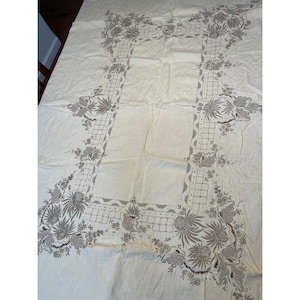 Pode incluir: Uma toalha de mesa retangular em tom off-white com bordados complexos em cinza escuro. O design inclui uma borda retangular central com um padrão de grade, acentuado por motivos detalhados de flores e folhas nos cantos e nas bordas.