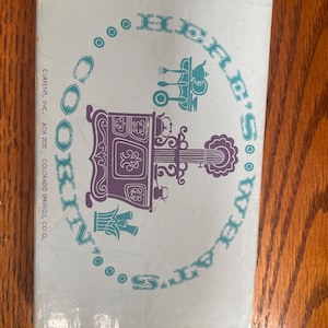 May include: A light blue rectangular box with the text "HERE'S COOKIN'" arranged in an oval. The center features a purple illustration of a vintage stove and a teal graphic of kitchen utensils, with "CURRENT, INC. BOX 2020 COLORADO SPRINGS, CO." printed on the left side.