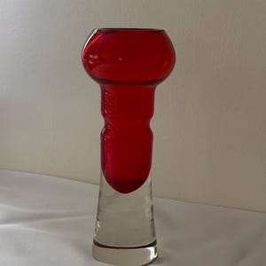 Pode incluir: Um vaso de vidro alto com uma secção superior em vermelho vibrante, apresentando um rebordo arredondado e bulboso, e um pescoço estreito que transita para uma base clara e transparente. O objeto tem uma superfície lisa e bicolor.