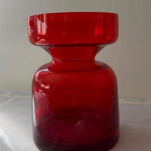 May include: A vibrant red glass vase featuring a wide, cylindrical top section that tapers into a narrow neck before expanding into a rounded, bulbous base. The translucent material allows light to pass through, highlighting the deep red color.
