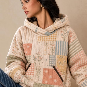 May include: A quilted hoodie featuring a patchwork design with various floral, gingham, and striped patterns in soft pastel shades of pink, yellow, blue, and green. The garment includes a hood, long sleeves, and two front pockets with dark zippers.