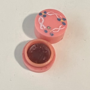 May include: A pink lip balm container with a floral design on the lid. The lip balm is a dark red color.