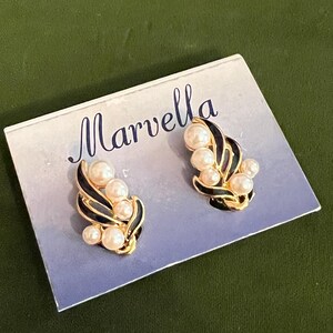 May include: A pair of gold-tone clip-on earrings with black enamel and faux pearls. The earrings are on a blue card with the word "Marvella" printed on it.