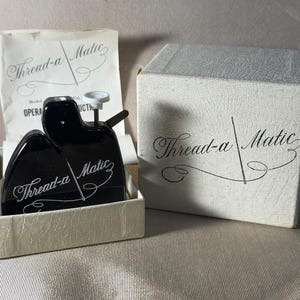 May include: A black Thread-a-Matic needle threader with a white plunger, housed in a textured white box. The device features the brand name in white script. Included is a white instruction sheet with the text Thread-a-Matic and Model OPERA.