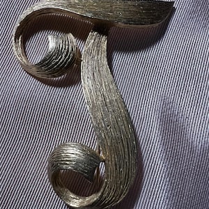 May include: A silver-toned brooch in the shape of the letter "J". The brooch has a textured, brushed metal finish. The design features a curved top and a flowing, stylized lower portion, with a pin on the back for attachment. The brooch is set against a gray background.