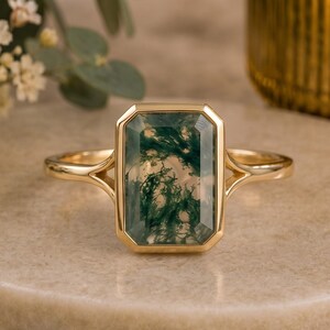 May include: A gold ring featuring a rectangular, emerald-cut moss agate gemstone with distinct green, fern-like inclusions. The stone is set in a smooth, polished gold bezel with a split shank design that tapers into a thin, rounded band.