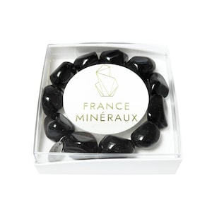 May include: A bracelet made of polished black tumbled stones arranged in a circle inside a square white box with a clear lid. A circular white label in the center features a gold geometric logo and the text FRANCE MINÉRAUX in gold lettering.