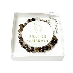 Puede incluir: Una pulsera hecha de piedras de cuarzo ahumado pulidas en una caja blanca. Una etiqueta circular blanca en el centro presenta un logotipo geométrico dorado y el texto FRANCE MINÉRAUX. La pulsera incluye una cadena de metal plateado con cierre.