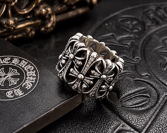 Op de afbeelding: Een brede zilveren ring met een herhalend patroon van verhoogde kruismotieven, geaccentueerd met kleine, heldere, sprankelende steentjes. De ring heeft een gepolijste metalen afwerking en diepe details tussen elk kruis voor een textuur.