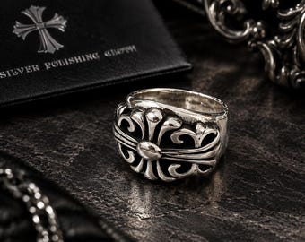 May include: A sterling silver ring featuring a prominent floral cross motif with intricate, raised scrollwork details. The band has a polished finish with darkened recessed areas to highlight the sculptural design elements.