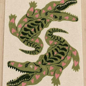 May include: A rectangular area rug with an off-white background featuring two green crocodiles. The crocodiles are decorated with dark green leaf-like patterns along their backs and small pink heart shapes across their bodies and limbs.