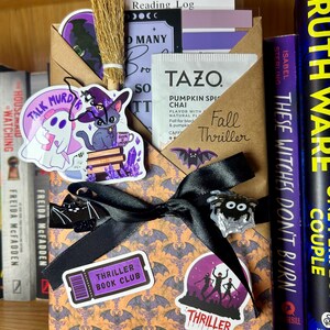 May include: A brown paper envelope decorated with a bat pattern, tied with a black ribbon, and adorned with stickers of a ghost, a cat, a spider, and a bat. It contains a Tazo Pumpkin Spice Chai tea bag, a reading log, and a miniature broom.