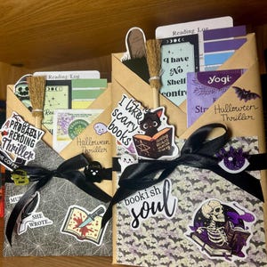 May include: Two brown paper envelopes decorated with Halloween-themed stickers, black ribbons, and miniature broomsticks. The envelopes contain reading logs, tea bags, and cards with text including "Probably Reading Thriller" and "Bookish Soul."