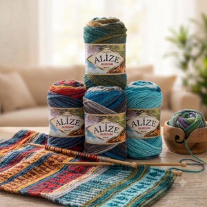 Peut inclure: Quatre pelotes de laine Alize Burcum Batik dans des tons de bleu, vert et rouge, empilées avec un bol à laine en bois contenant une petite pelote. Un tricot à rayures multicolores est posé devant avec des aiguilles à tricoter en bois.