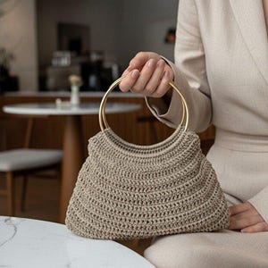 May include: A beige crochet handbag featuring a structured, trapezoidal body with a textured, ribbed stitch pattern. The bag is equipped with two circular, polished gold-tone metal ring handles that provide a firm grip for carrying the accessory.