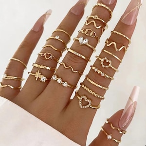 Peut inclure: Une collection de bagues dorées portées sur les doigts, présentant divers designs incluant des formes de cœur, des bandes ondulées, des motifs d'étoiles, des textures torsadées et de petites pierres claires. Les bagues sont empilées sur plusieurs doigts, mettant en valeur une variété de fines bandes métalliques avec des finitions polies et des éléments décoratifs.