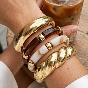 Peut inclure: Un empilement de quatre bracelets comprenant un jonc doré lisse, un bracelet à maillons en résine marron foncé avec des détails dorés, un bracelet à maillons en résine marbrée blanc et marron avec des détails dorés et un jonc torsadé doré.