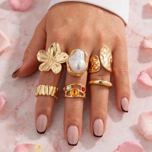 Peut inclure: Un ensemble de six bagues dorées portées sur les doigts. Les bagues présentent des designs variés, dont une grande fleur, une pierre ovale blanche, une bande texturée, une bande ondulée, une bande avec des pierres colorées et une bague lisse.