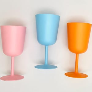 May include: Three solid-coloured plastic wine glasses in pastel pink, light blue, and bright orange. Each glass features a smooth, matte finish, a rounded bowl, a slender stem, and a wide circular base, standing upright in a simple arrangement.