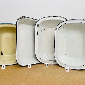 May include: Four rectangular enamel basins arranged in a row, labelled with small white tags numbered one through four. The first basin is cream-coloured with a blue rim, while the others are white with black rims, all showing signs of wear and rust.
