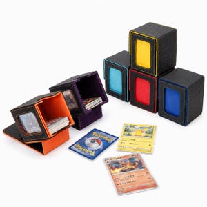 Può includere: Una collezione di scatole per mazzi nere testurizzate con rivestimenti interni colorati e finestre di visualizzazione frontali. Le scatole sono disposte in pila e in configurazione aperta, accompagnate da tre carte collezionabili con Pikachu, Charizard e un retro blu.