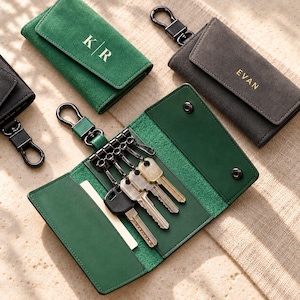 May include: A collection of suede leather key cases in green and gray, featuring metal snap closures and key hooks. One green case is open, revealing internal slots and four metal keys. Personalization includes the letters K|R and the name EVAN.