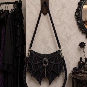 May include: A black crocheted handbag shaped like bat wings, featuring a long braided shoulder strap and a central oval black gemstone pendant set in an ornate bronze-toned metal frame with a small dangling bead accent at the bottom.
