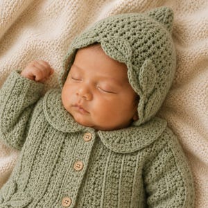May include: A sage green crocheted long-sleeve cardigan with a rounded collar and three light brown wooden buttons, paired with a matching pointed crocheted bonnet featuring decorative leaf-shaped accents along the front edge.