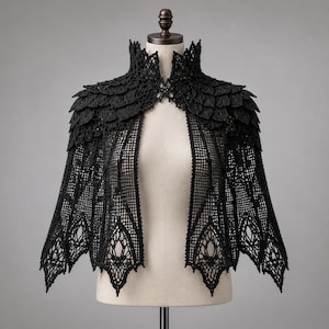 Peut inclure: Un boléro cape en crochet noir doté d'un col montant et de détails complexes en forme de feuilles superposées sur les épaules. Le vêtement présente un corps à motif quadrillé qui se termine par un ourlet festonné ajouré et pointu.