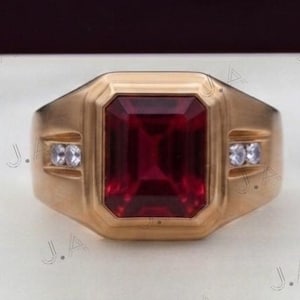 Puede incluir: Un anillo de oro con una gran piedra preciosa de color rojo intenso y corte esmeralda rectangular en el centro. La banda incluye dos pequeñas piedras de acento redondas y transparentes a cada lado del engaste central.