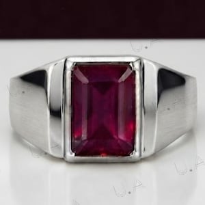 Op de afbeelding: Een zilverkleurige ring met een grote, rechthoekige, dieprode edelsteen gezet in een gladde, gepolijste metalen band. De steen heeft een geslepen facetvorm die het licht reflecteert en de band heeft een breed, solide ontwerp dat taps toeloopt.