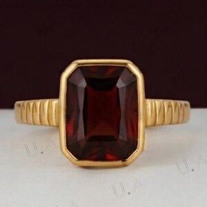 Puede incluir: Un anillo de oro con una gran piedra preciosa rectangular de color rojo intenso en un engaste de bisel. La banda del anillo presenta una textura distintiva de estrías o ranuras verticales en los lados, que se extiende desde el engaste central.
