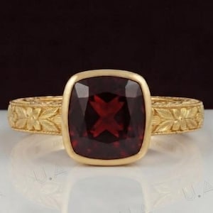 Puede incluir: Un anillo de oro con una gran piedra preciosa de granate rojo oscuro, de corte cojín, engastada en un bisel liso. La banda está hecha de oro amarillo y muestra un intrincado patrón floral en relieve que rodea toda la circunferencia.