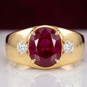 May include: A gold ring featuring a large, oval-cut, deep red gemstone at the centre, flanked by two small, round, clear, brilliant-cut diamonds. The band is smooth, polished yellow gold with a wide, tapered design that holds the stones securely.