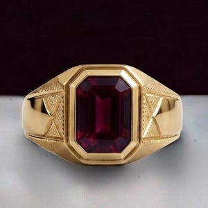 May include: A gold ring featuring a large, rectangular, deep red gemstone set in the centre. The band is crafted from polished gold and displays intricate, textured geometric patterns on both sides of the central stone setting.
