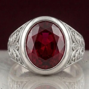 May include: A silver ring featuring a large, oval-cut, deep red gemstone set in the centre. The band of the ring is crafted with intricate, embossed scrollwork and leaf-like patterns that extend along the sides of the metal setting.
