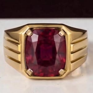 Puede incluir: Un anillo de oro con una gran piedra preciosa de color rojo intenso y talla cojín en el centro. El aro del anillo tiene un acabado pulido con tres ranuras horizontales a cada lado, que conducen al engaste central que sujeta la piedra.