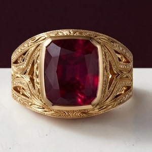 Op de afbeelding: Een gouden ring met een grote, rechthoekige, kussengeslepen dieprode edelsteen in het midden. De band is gemaakt van geelgoud en vertoont ingewikkelde, gegraveerde krulpatronen over het gehele oppervlak van de metalen zetting.