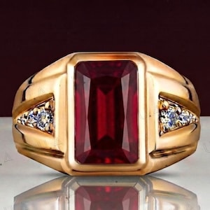 Op de afbeelding: Een gouden ring met een grote, rechthoekige, donkerrode edelsteen in het midden. De ringband is breed en gepolijst, met twee driehoekige zijaccenten, elk met drie kleine, heldere, fonkelende rondgeslepen stenen in het metaal gezet.