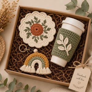 May include: A gift box containing a round cream-colored crochet coaster with a rust and green leaf pattern, a green crochet cup sleeve with a cream leaf applique, a rainbow-shaped crochet keychain, and a tag reading Handmade with love.