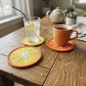 May include: Three round crochet coasters designed to look like citrus fruit slices. One coaster resembles a yellow lemon slice, while two others feature orange and white patterns. These circular textile items are placed under a glass and a mug.