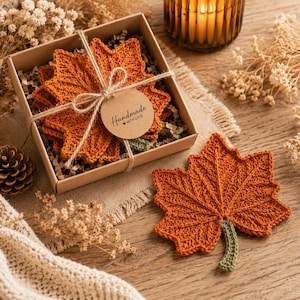 May include: A set of orange crochet leaf coasters in a square cardboard box tied with twine and a round tag reading Handmade with Love. One matching orange crochet leaf coaster with a green stem is placed outside the box on a wooden surface.