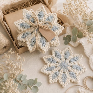 May include: A set of star-shaped crochet coasters featuring a cream and light blue colour pattern. One coaster is placed outside a cardboard gift box, while a stack of identical coasters is tied together with twine and a blank brown paper tag.
