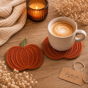 Peut inclure: Deux dessous de verre au crochet en forme de citrouille orange avec des tiges vertes. L'un des dessous de verre est placé sous une tasse en céramique mouchetée remplie de café. Une étiquette rectangulaire marron porte l'inscription Handmade with love.