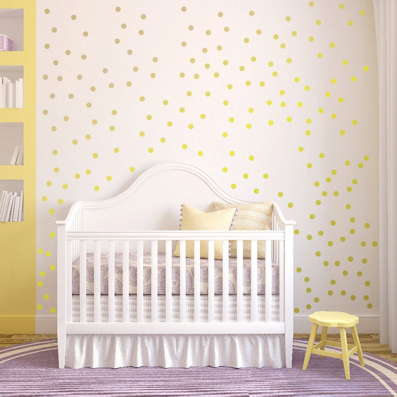 May include: A white crib with a white ruffled skirt and a yellow polka dot wall decal. The crib is in a nursery with a yellow bookshelf and a purple rug.
