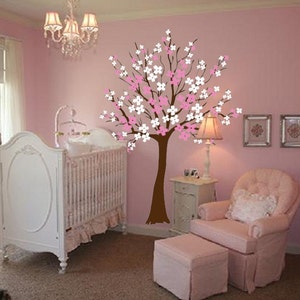 May include: A pink and white floral wall decal of a tree with a brown trunk. The decal is in a nursery with a white crib, a pink armchair, and a pink ottoman.
