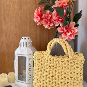 May include: A rectangular handbag crafted from thick, pale yellow yarn in a repeating star-like crochet pattern. The bag features two sturdy, rounded handles made of the same yellow yarn, providing a structured shape for this handmade accessory.